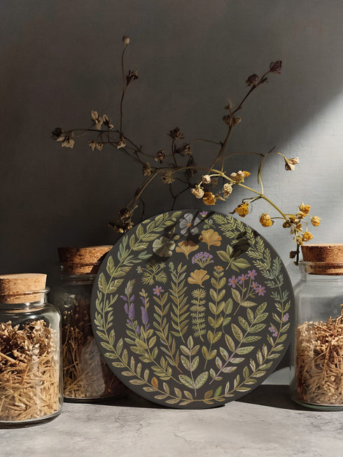 Circular ceramic coaster with black background featuring intricate botanical illustrations of various herbs, flowers and leaves in green, purple and gold colors with white ceramic edge and cork backing.